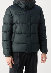 Uomo che indossa un giubbotto puffo nero con tasche con zip e bottoni a pressione, abbinato a pantaloni neri larghi su uno sfondo neutro.
