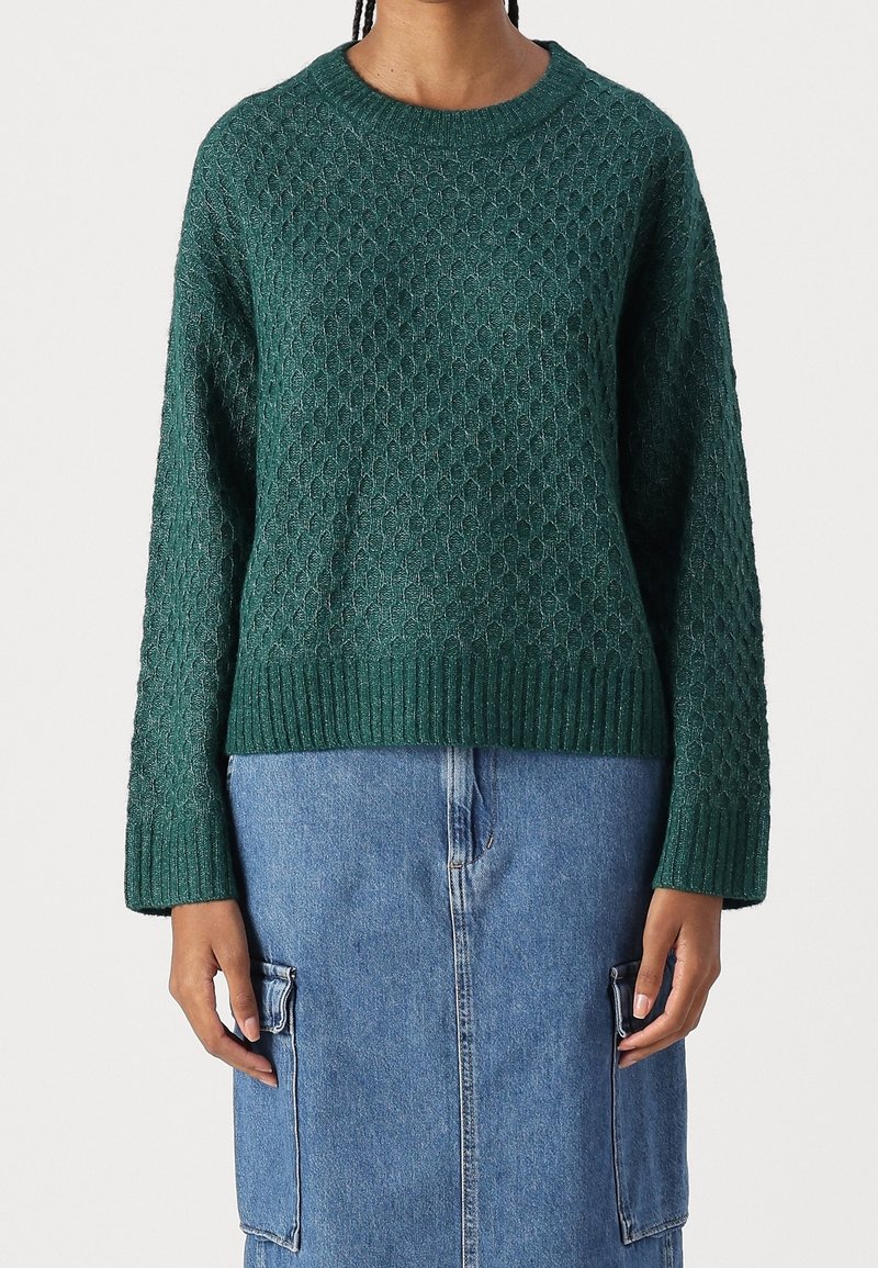 Personne portant un pull en maille texturée vert foncé et une jupe en denim bleu avec de grandes poches latérales, se tenant devant un fond uni.