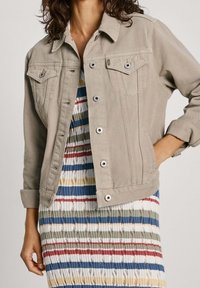 Chaqueta de mezclilla beige con un corte clásico, que cuenta con dos bolsillos en el pecho, botones plateados y mangas enrolladas, combinada con un vestido a rayas debajo.