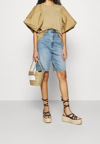 Khaki oversized košile s balónovými rukávy, modré džíny šortky, černé sandály na platformě s pásky a pletená kabelka.