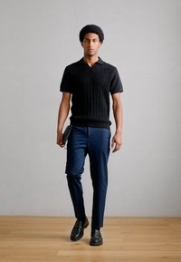 Μαύρο πλεκτό πόλο πουκάμισο με κοντά μανίκια, συνδυασμένο με ναυτικό μπλε παντελόνι. Το μοντέλο στέκεται σε ξύλινο πάτωμα, κρατώντας ένα μαύρο τάμπλετ.