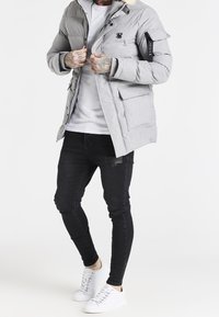 Graue gepolsterte Jacke mit abnehmbarem Kapuzenkragen, ausgestattet mit einem kleinen Logo, vier Fronttaschen und kontrastierenden schwarzen Ärmeldetails. Kombiniert mit schwarzen Jeans und weißen Sneakern.