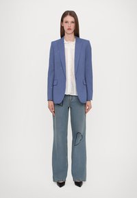 Blauwe blazer met subtiele versieringen, witte blouse met ruffles, versleten wijde spijkerbroek en zwarte hakken.