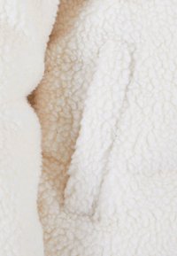 Tissu en polaire blanche avec une surface douce et texturée. Comprend une poche cachée, avec une doublure beige légèrement plus claire visible en dessous.