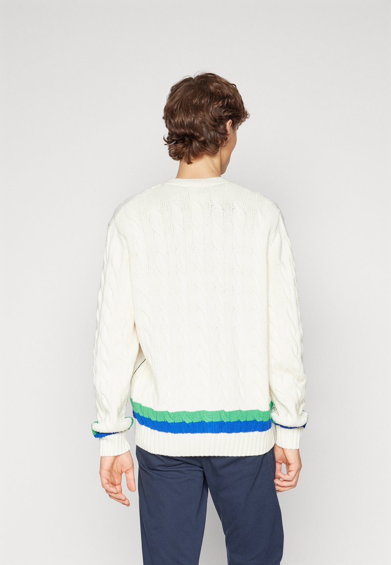 Pull en maille torsadée crème avec un col rond et des poignets côtelés, présentant un ourlet à motif bicolore en vert et bleu.