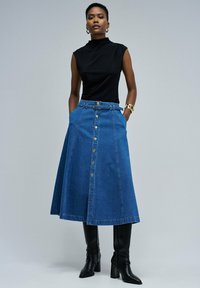 Kvinna står med händerna i fickorna och bär en ärmlös svart topp, en blå knäppt denim-midi kjol med bälte, och svarta knähöga stövlar.
