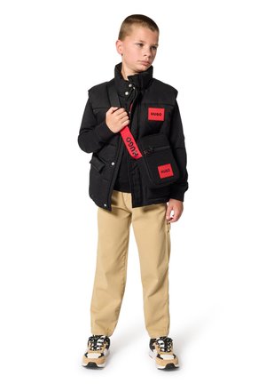 Doudoune sans manches noire avec des écussons logo rouges, chemise noire à manches longues, pantalon beige et baskets. Comprend un sac bandoulière noir avec une sangle rouge.