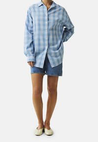 Modrá a bílá kostkovaná košile s dlouhými rukávy, nošená přes vybledlé džíny šortky. Model má na sobě světlé ploché boty.