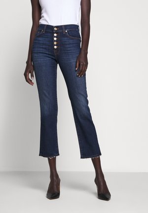 Femme portant un jean court taille haute bleu marine avec des ourlets effilochés et une fermeture à boutons dorés, associé à des talons pointus noirs et un haut blanc.
