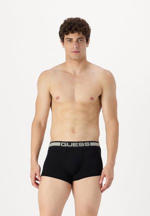 Des boxers en coton noir disposent d'une ceinture élastique grise avec la marque "GUESS". Le design est ajusté et arrive au milieu de la cuisse.