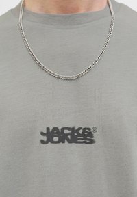 Gray cotton t-shirt featuring a black logo on the front. The shirt has a crew neck and is paired with a silver chain necklace.