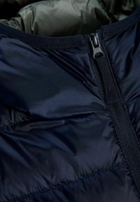 Gros plan sur le col d'une doudoune noire avec une fermeture éclair partiellement fermée et des détails de couture visibles.
