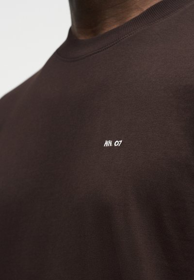 Κοντινό πλάνο ενός ατόμου που φοράει μια σκούρα καφέ μπλούζα με στρογγυλή λαιμόκοψη και λευκό κεντητό κείμενο "NN. 07" στο αριστερό μέρος του στήθους.