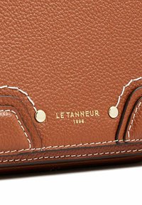Prodotto in pelle marrone con texture, caratterizzato da accenti in oro, cuciture bianche e logo impresso "LE TANNEUR 1898" sulla superficie.
