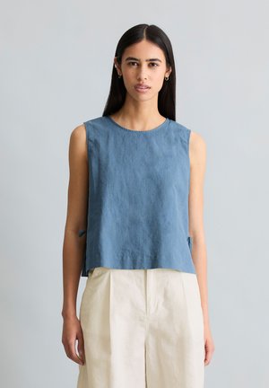 Femme aux longs cheveux foncés portant un haut bleu sans manches et un pantalon blanc taille haute, debout devant un fond gris clair.