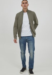 Giacca verde oliva con zip, t-shirt bianca, jeans blu e sneakers nere. La giacca presenta un colletto alto e polsini a costine.