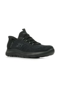 Zapatilla negra de Skechers tipo slip-on con parte superior de malla, cordones elásticos y suela acolchada, inclinada a la izquierda sobre un fondo blanco.