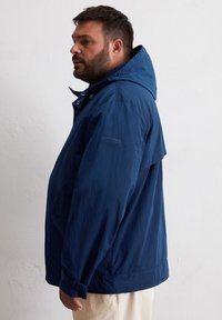 Marineblaue leichte Jacke mit Kapuze, ausgestattet mit Druckknöpfen, einem strukturierten Stoff und einer Seitentasche. Logo auf dem linken Ärmel.