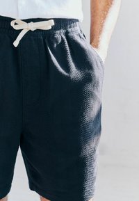 Pantalones cortos azul marino con una tela texturizada, cinturilla elástica y un cordón beige. Cuenta con bolsillos laterales y un ajuste relajado.