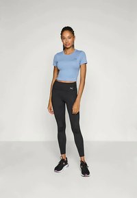 Svetlo modra kratka majica in črne legice s Puma logotipom. Kratka zasnova je oprijeta; legice so visoko pasne in raztegljive. Črne superge dopolnjujejo videz.