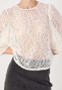 Blouse en dentelle crème avec manches courtes bouffantes, ornée de motifs floraux complexes. Portée sur une jupe ajustée gris foncé avec des accents subtils.