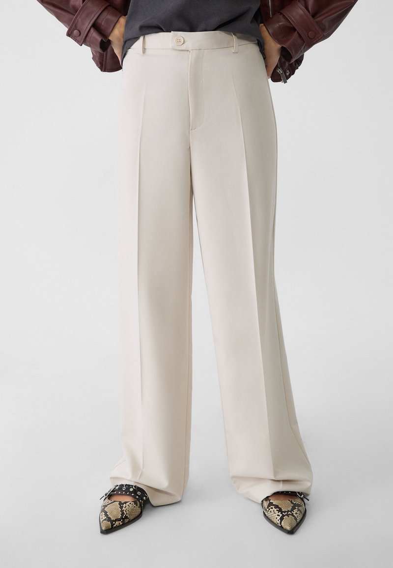Personne portant un pantalon large crème taille haute, des chaussures pointues en cuir de serpent noir avec des brides, et un haut sombre avec une veste en cuir marron.