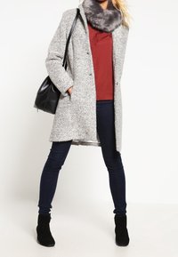 Lichtgrijze, textuurwoljas met een nepbontkraag, gecombineerd met donkere jeans en zwarte enkelboots. Model draagt een zwarte handtas.