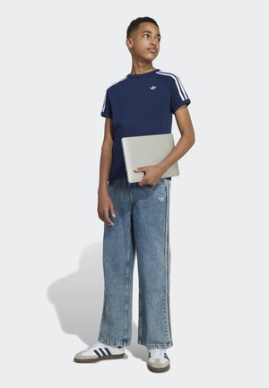 Teenager v modrém tričku Adidas, širokých džínách a bílých teniskách drží zavřený laptop a dívá se mírně doprava.