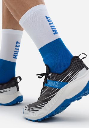 Jambes portant des chaussettes blanches et bleues de la marque Millet et des chaussures de course Millet noir, blanc et bleu avec des semelles texturées sur un fond clair.