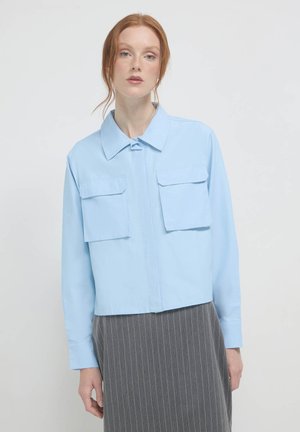 Himmelblaues, cropped Hemd mit einem spitzen Kragen, das zwei Fronttaschen und lange Ärmel hat. Glatte Textur, Knopfverschluss am Hals.