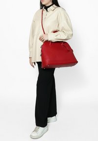 Rode leren handtas met een afgeronde vorm, twee handvatten bovenaan en een verstelbare schouderriem; heeft zilverkleurige hardware en stiksels als accent.