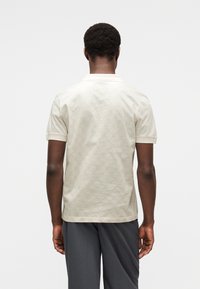 Ανοιχτό μπεζ πόλο πουκάμισο με διακριτικό γεωμετρικό σχέδιο. Κοντά μανίκια και ίσιος γιακάς. Μαλακή υφή, απλός σχεδια σμός και εφαρμοστή γραμμή.