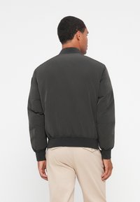 Giubbotto bomber nero con una texture liscia, polsini e fascia in vita a coste, dotato di colletto alto e maniche leggermente imbottite.