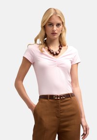 Jasnoróżowa bluzka z krótkim rękawem i dekoltem w serek z marszczeniami, zestawiona z brązowymi spodniami w wysokim talii i masywną, ciemną biżuterią.