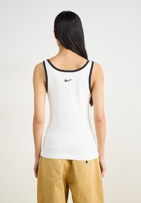 White sleeveless tank top with black trim and a small Nike logo on the back, paired with beige baggy pants. Smooth cotton fabric.