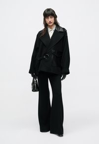 Žena oblečená v čiernych nohaviciach s širokými nohavicami, nadrozmernej čiernej bunde s koženými detailmi, čiernych rukaviciach a držiaca čiernu kabelku, na bielom pozadí.