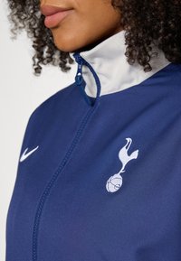 Blå zip-up jacka med vit krage, med en broderad Tottenham-logotyp. Tillverkad av slätt tyg, utformad för atletisk användning.