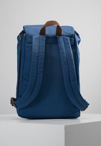 Zaino blu con dettagli in pelle marrone, forma rettangolare, materiale in tessuto, spallacci imbottiti e chiusura con zip.
