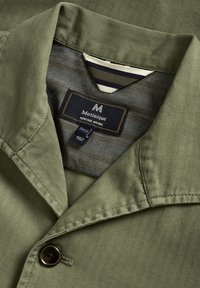 Olivgrüne Jacke mit Fischgrätstruktur, ausgestattet mit einem Reverskragen, einem einzelnen Knopfverschluss und einem Etikett mit der Aufschrift "Matiniqué Heritage Apparel."