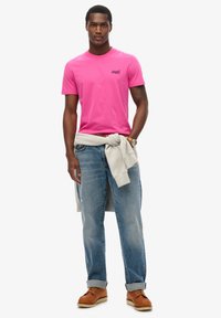 T-shirt di cotone rosa, con collo a girocollo, presente un piccolo logo. Abbinato a jeans azzurri chiari e un felpa grigia annodata in vita. Scarpe marroni.
