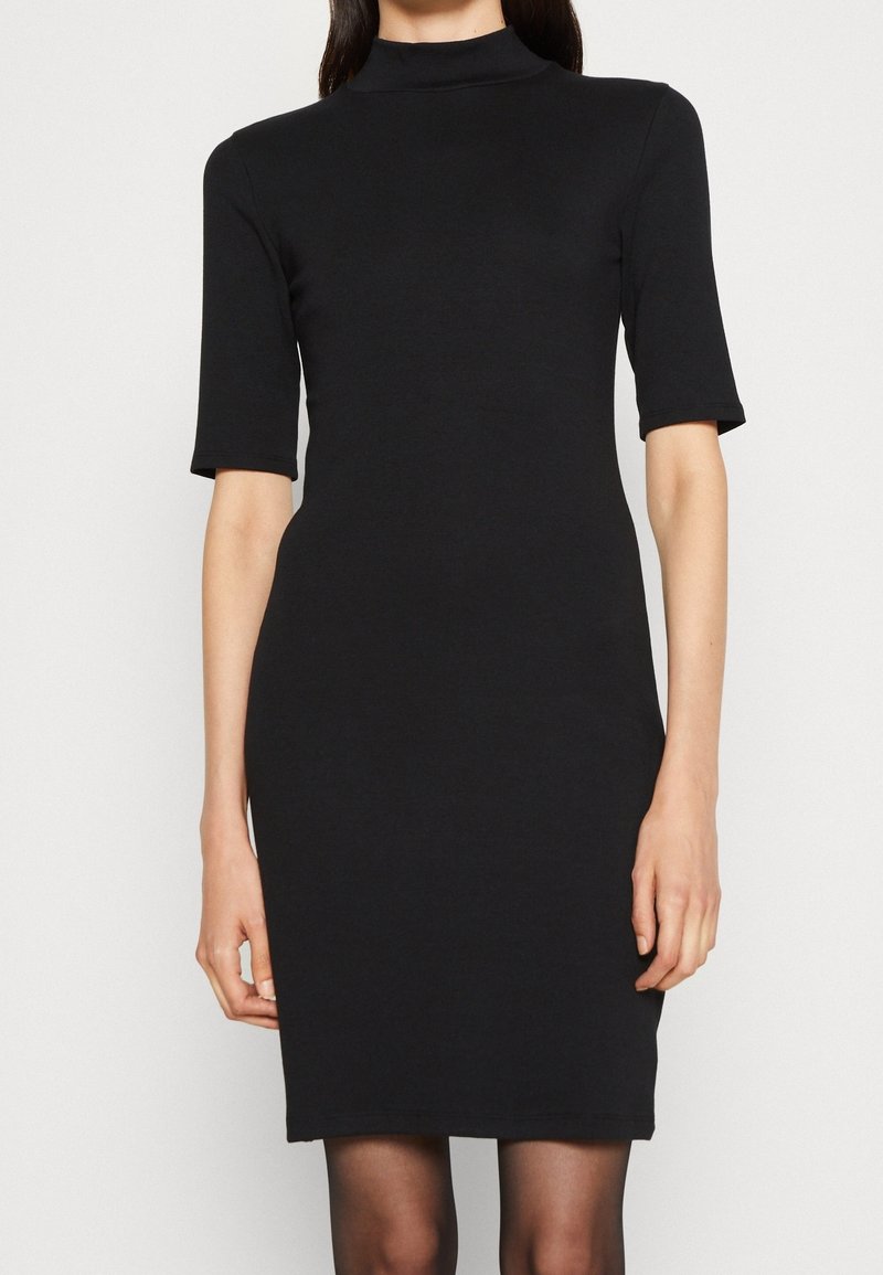 Femme portant une robe noire cintrée avec un col haut et des manches aux coudes, debout devant un fond clair uni.