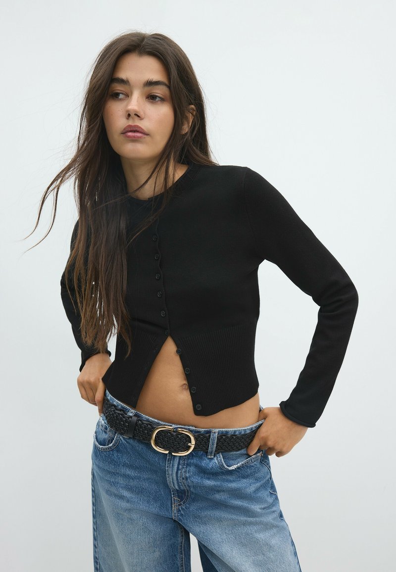 Jeune femme aux longs cheveux foncés portant un cardigan noir boutonné et un jean bleu avec une ceinture tissée noire, posant devant un fond clair uni.