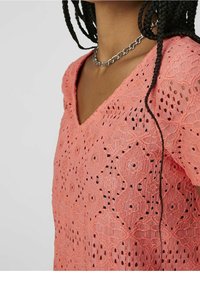 Haut en dentelle rose avec un décolleté en V et un motif intricate avec des découpes. Comprend un collier en chaîne argenté. La texture est légère et aérienne.