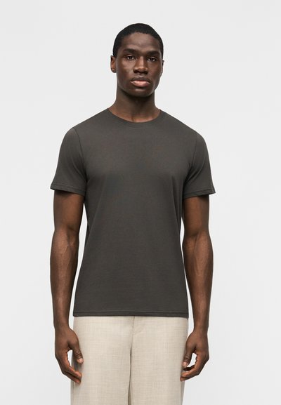 Homme debout devant un fond blanc, portant un t-shirt gris foncé uni à manches courtes et un pantalon beige clair, faisant face à l'avant avec une posture détendue.