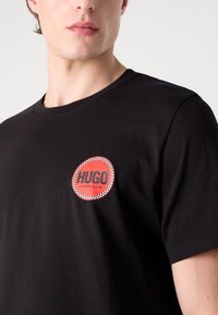 Black cotton T-shirt featuring a red circular logo with checkered border reading "HUGO WINNING BLEND" on the left chest area.