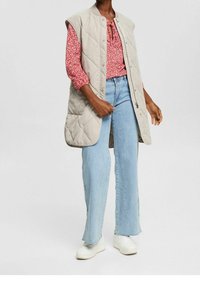 Gilet lungo beige trapuntato con colletto alto, chiusura a bottoni, abbinato a una blouse floreale rossa e a jeans a gamba larga di un azzurro chiaro. Sneakers bianche.
