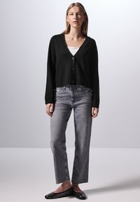 Svart, kortärmad cardigan med V-ringning och knappstängning, buren över en vit linne. Kombinerad med grå raka jeans och svarta platta skor.
