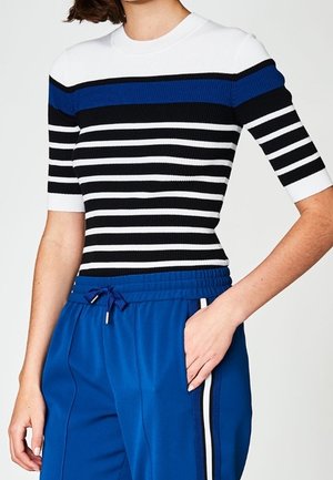 Femme portant un haut à manches courtes rayé blanc, bleu et noir, associé à un pantalon de sport bleu avec une bande latérale blanche et noire.