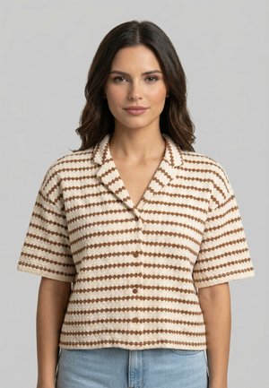 Mujer de cabello oscuro y longitud media, lleva una blusa de botones de manga corta a rayas beige y marrón, y jeans azul claro, de pie contra un fondo gris.