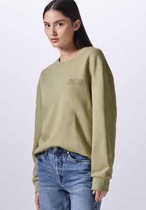 Sweatshirt oversize verde oliva con polsini e scollatura a costine, caratterizzato da un piccolo logo stampato sul lato sinistro del petto. Indossato con jeans blu.